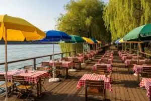 Terrasse ensoleillée d'une guinguette parisienne au bord de l'eau, tables à nappes vichy et parasols colorés, saules pleureurs